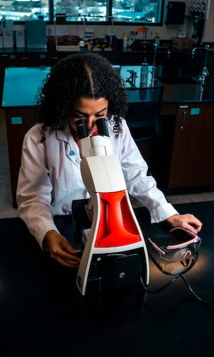 Student in a lab coat examines a specimen through a compound microscope with illuminated base at a laboratory bench, with safety goggles and smartphone nearby.