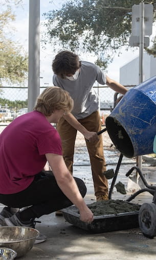 Students mix and pour concrete from a rotating drum cement mixer into a tray.