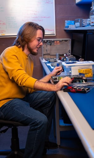 Student assembles and tests a printed circuit board with electronic components and tools at an electronics lab workbench, with lab instruments and a whiteboard in the background.