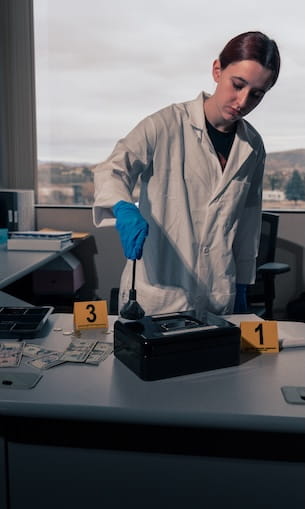 Student in a lab coat and nitrile gloves dusts a device for fingerprints beside numbered evidence markers on a laboratory table, simulating a forensic investigation.