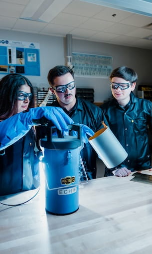 Three students wearing safety glasses and gloves operate a cryogenic materials container labeled ECH2, pouring liquid into the insulated vessel during a laboratory experiment.
