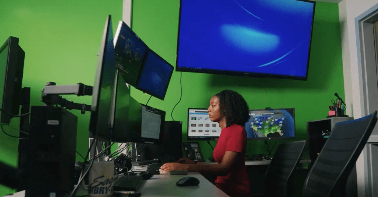 Student monitors multiple weather and radar displays at a workstation with several mounted screens and wall monitors in a meteorology lab with green screen backdrop.