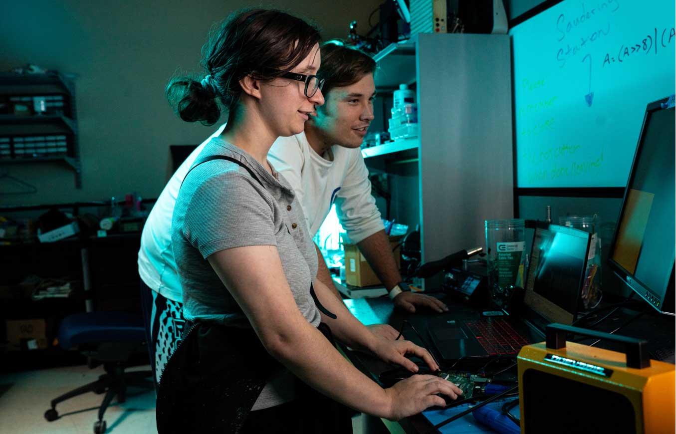 Two students collaborate at a lab workstation, programming and testing a microcontroller board connected to a laptop and external monitor, with electronics equipment and a whiteboard in the background.