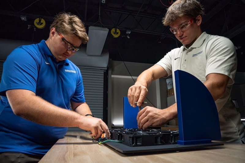 Two students wearing safety glasses assemble and wire an engineering test fixture with blue vertical panels and embedded electronics on a laboratory workbench.