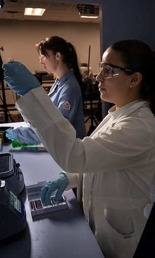 Students wearing lab coats, safety glasses, and gloves prepare samples with pipettes and a benchtop centrifuge in a biomedical laboratory.