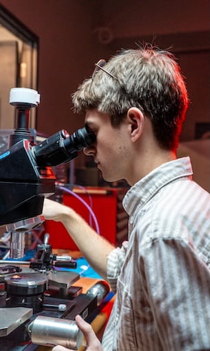 Student peers into a compound light microscope with mechanical stage and objective turret, adjusting the focus knobs in a laboratory workspace with red task lighting.
