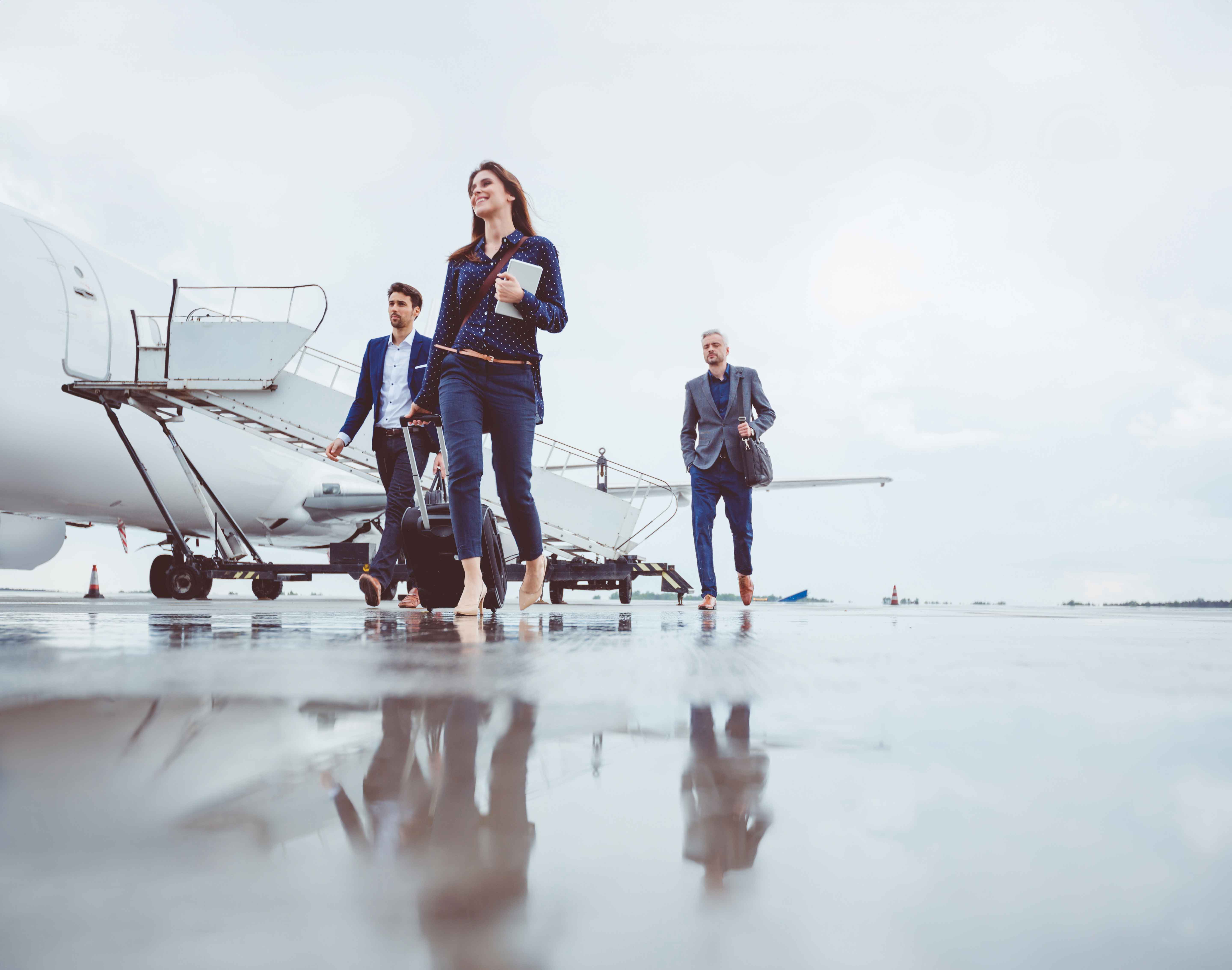 Three business professionals walk across an airport ramp beside a parked passenger jet and mobile boarding stairs, carrying luggage and a briefcase on a wet tarmac.