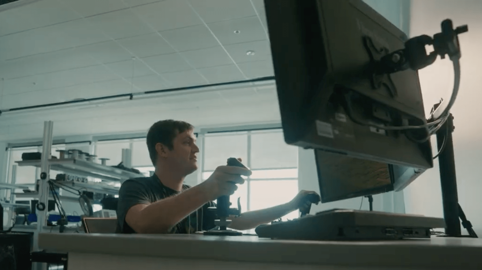 Student operates dual flight control joysticks at a desktop simulator station with mounted monitors in an aerospace laboratory.