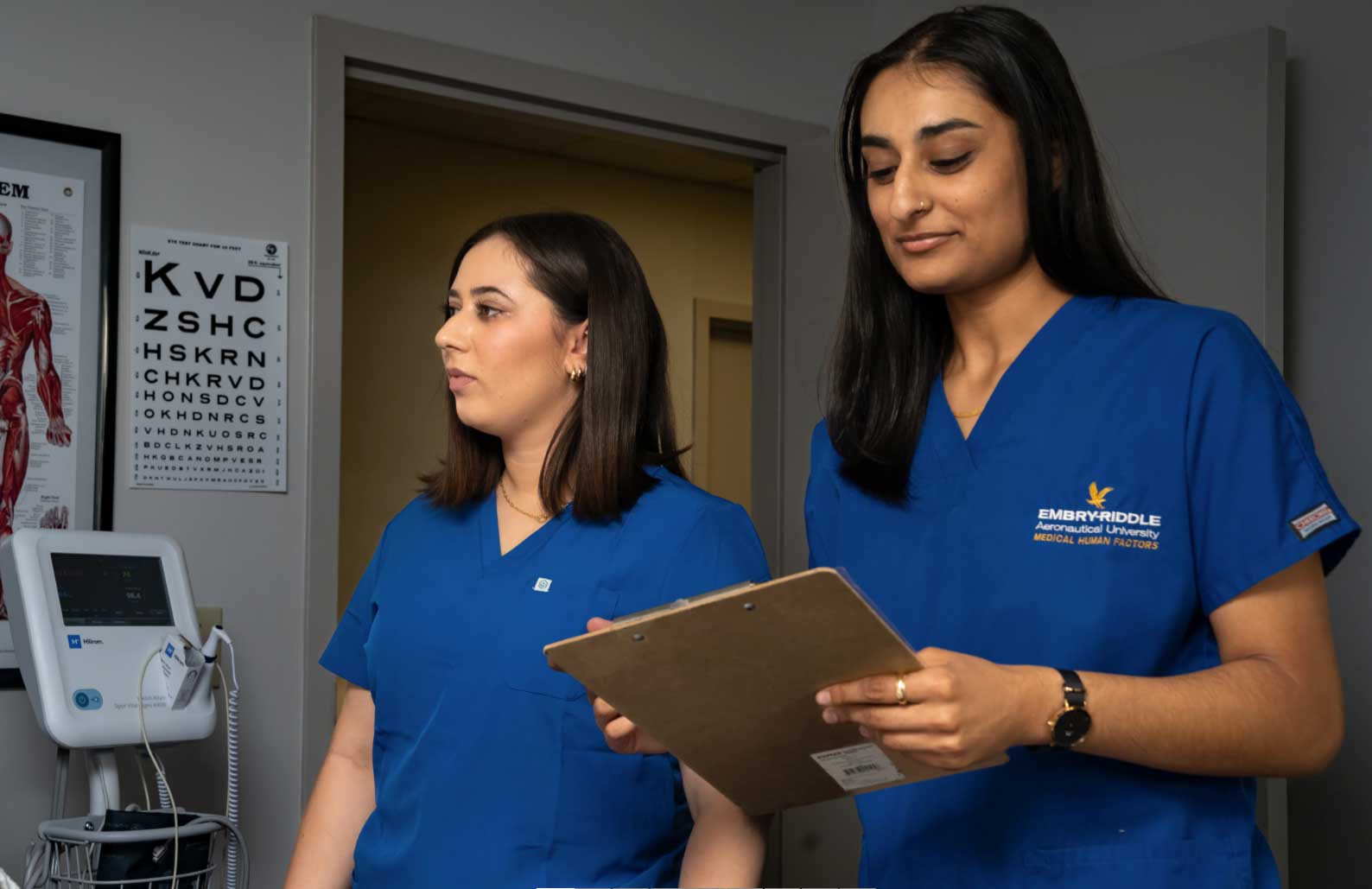 Two students in Embry-Riddle Medical Human Factors scrubs review a patient chart on a clipboard beside a vital signs monitor and eye chart in a clinical lab setting.