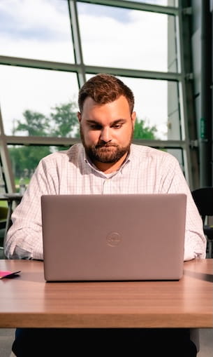 Student works on a laptop at a table inside a campus building with large glass windows and trees visible outside.