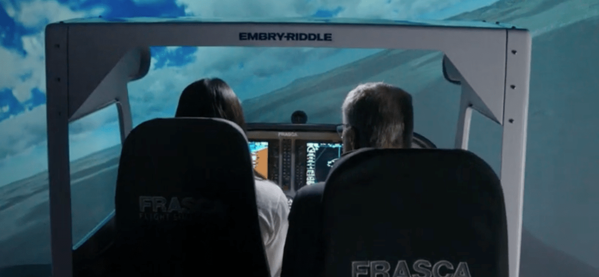 Two students seated in a FRASCA flight simulator cockpit beneath an Embry-Riddle frame, viewing glass cockpit avionics displays and projected outside flight scenery.