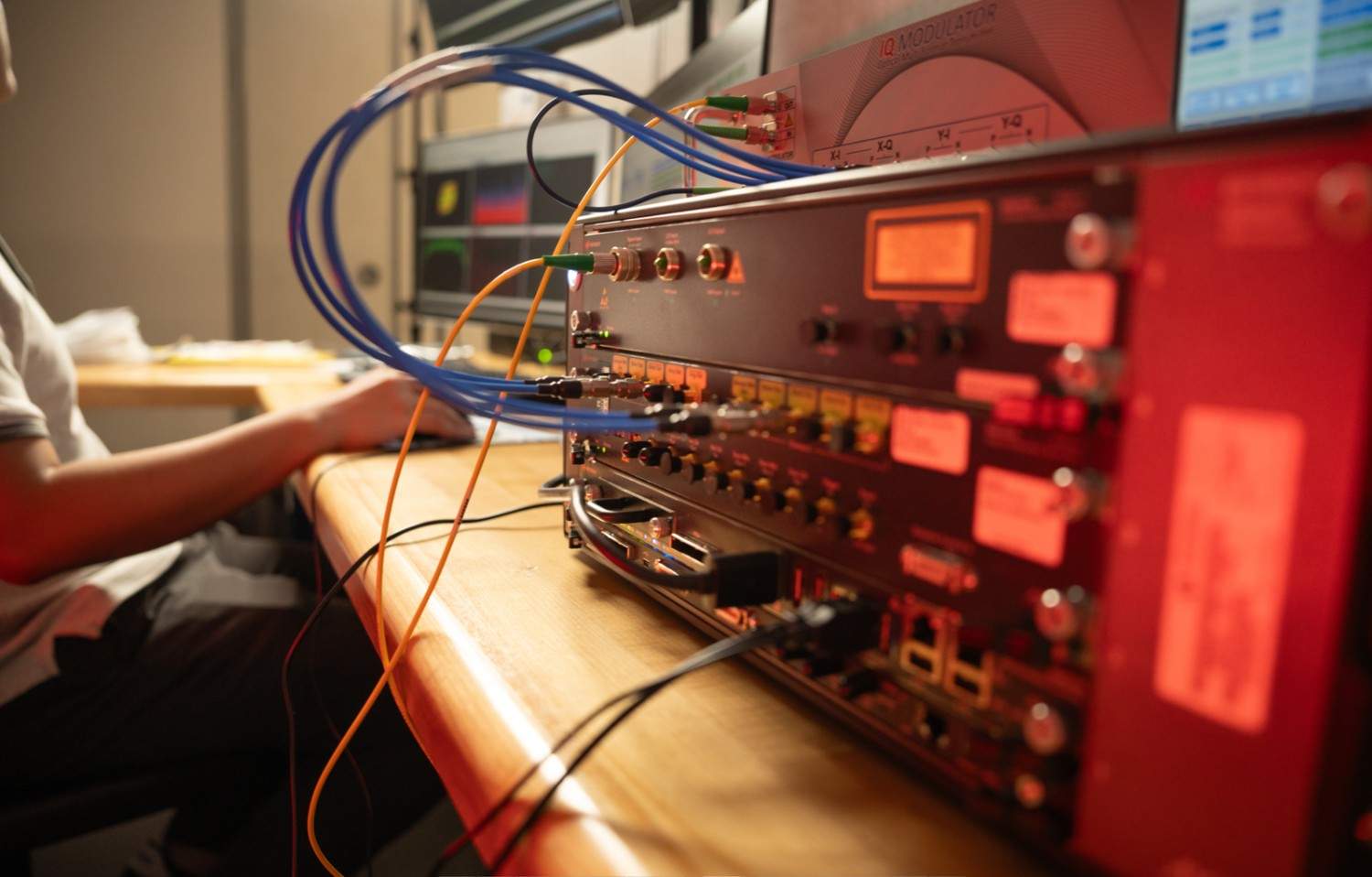 Close view of an IQ modulator and RF signal processing equipment with coaxial connectors, fiber optic cables, and control interfaces on a laboratory bench, as a student operates a computer nearby.