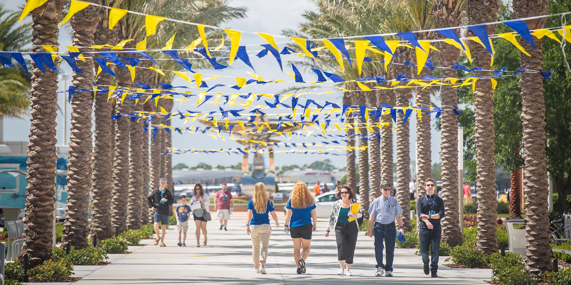 Students and family walk through rows of palm trees, along the Daytona Beach Campus promenade, which is draped with yellow and blue flag garland.