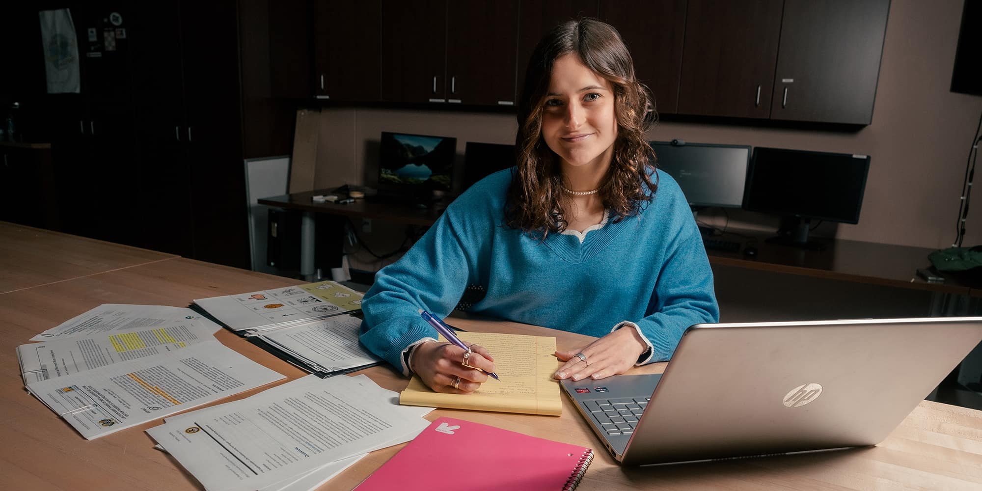 Alexandra Houston, wearing a blue sweater and pearl necklace, sits at a table in a classroom with a laptop, notepad and printed journal articles.