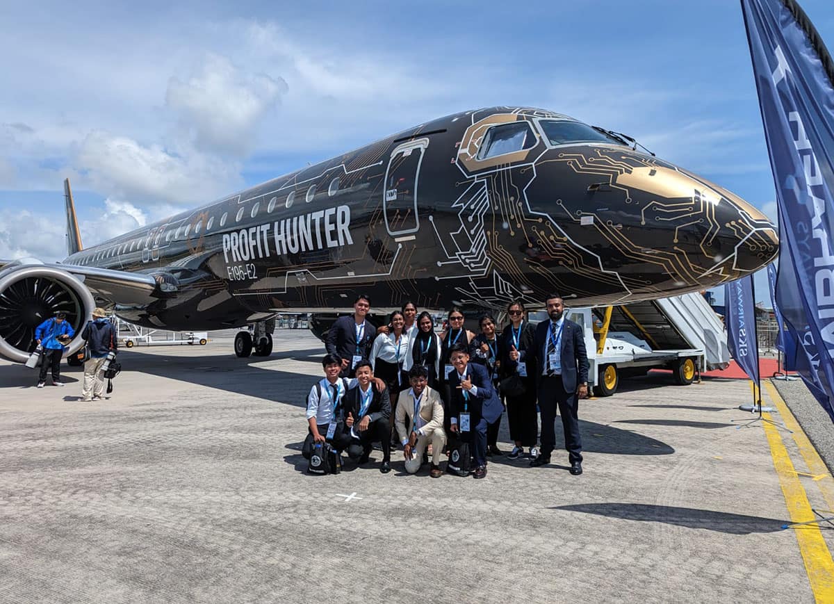 Arjun poses with a dozen others in front of a black jet with bronze and white microchip circuitry patterns. Lettering on the side reads, Profit Hunter.