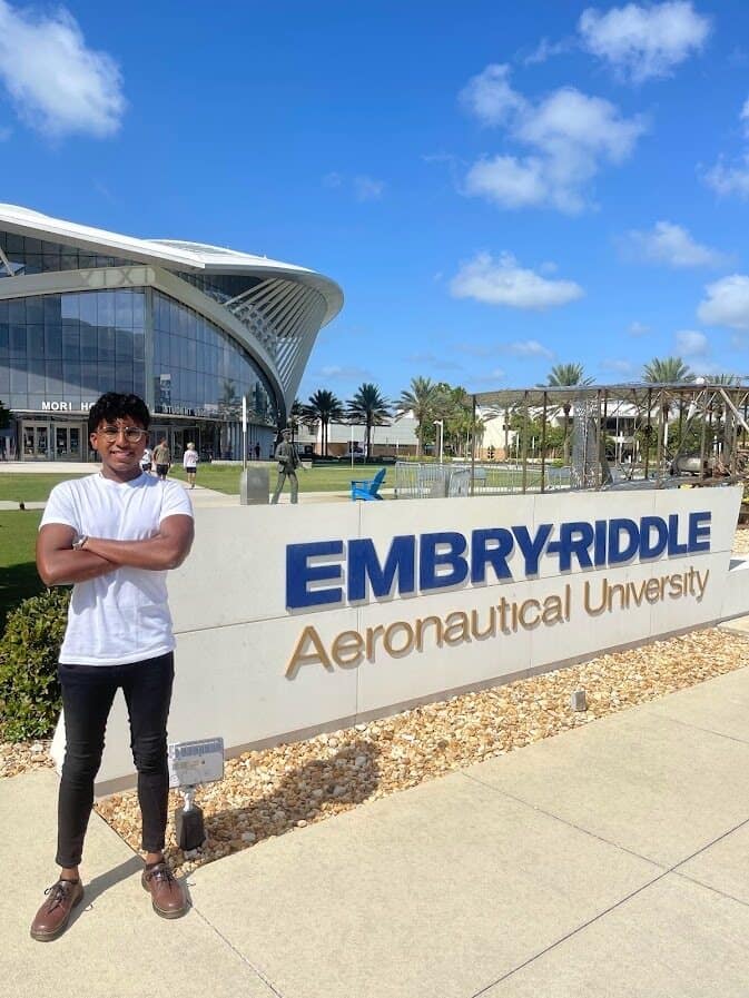 Arjun, in a white t-shirt, poses next to the Embry-Riddle monument sign on the Daytona Beach Campus on a sunny day.