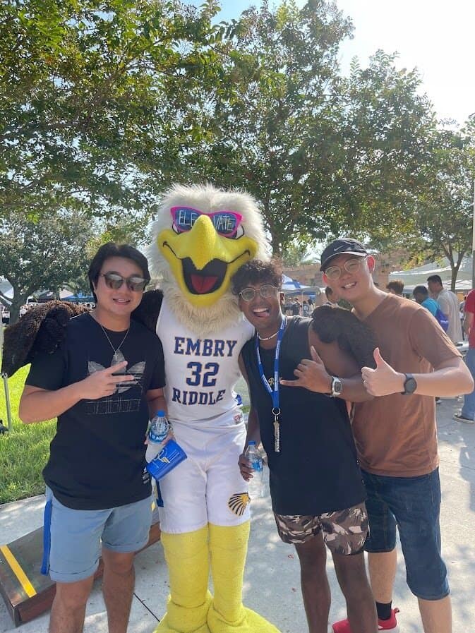 Arjun, wearing a t-shirt and camouflage shorts, makes a hang ten sign. He poses with two other students and the Embry-Riddle eagle mascot, who is wearing sunglasses and a basketball jersey.