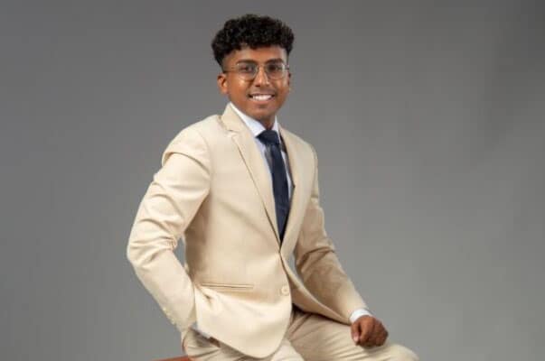 Arjun, a man with medium-dark skin tone, poses against a grey backdrop, wearing a cream-colored suit and navy blue tie and smiling broadly.
