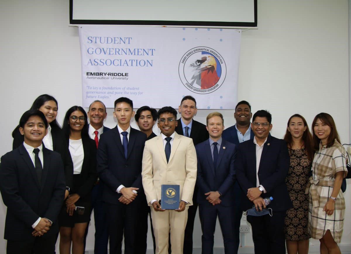 Arjun, in a cream-colored suit, poses with several other men and women with a wide variety of apparent ethnicities under a poster that reads Student Government Association.
