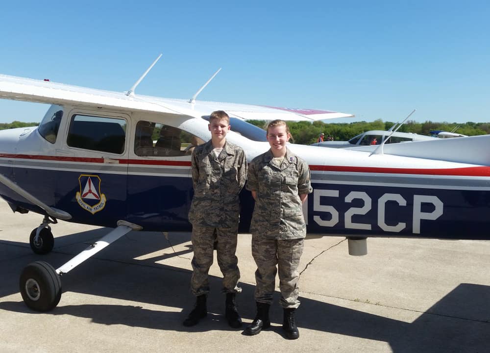 Calen and a young man both wear camouflage fatigues and stand in front of a prop plane with an insignia that reads Civil Air Patrol.