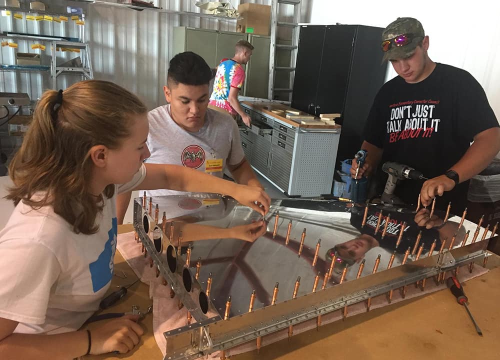 Calen - hair pulled back, wearing a t-shirt - and two other students add pins to a reflective metal plane part.