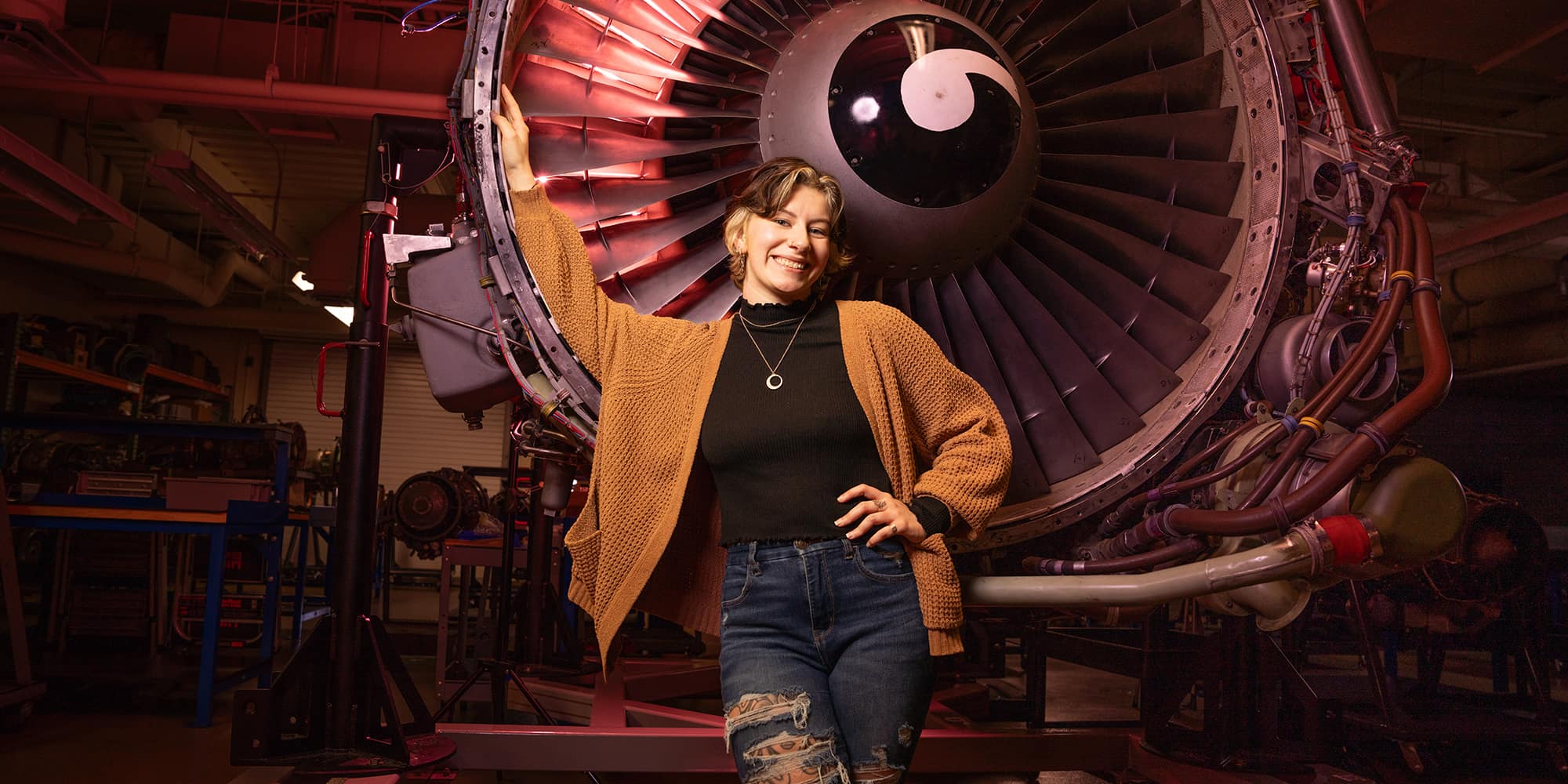 Calen, a woman with light skin tone and short, two-toned hair, stands in a maintenance shop in front of a large engine fan.