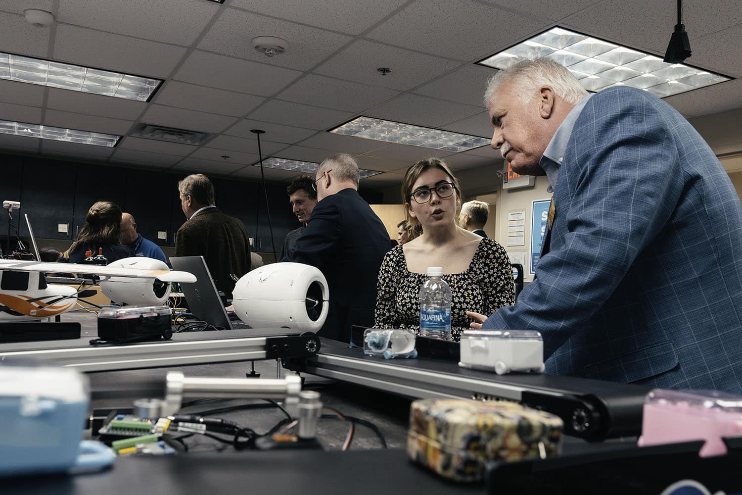 A woman with blonde hair and glasses talks to a man with a white mustache and hair near a table with various computer equipment, including a large drone. Several other people mill about in the background.
