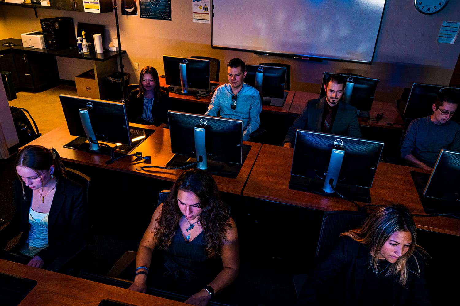 Several students sit in front of desktop computers in a darkened classroom.