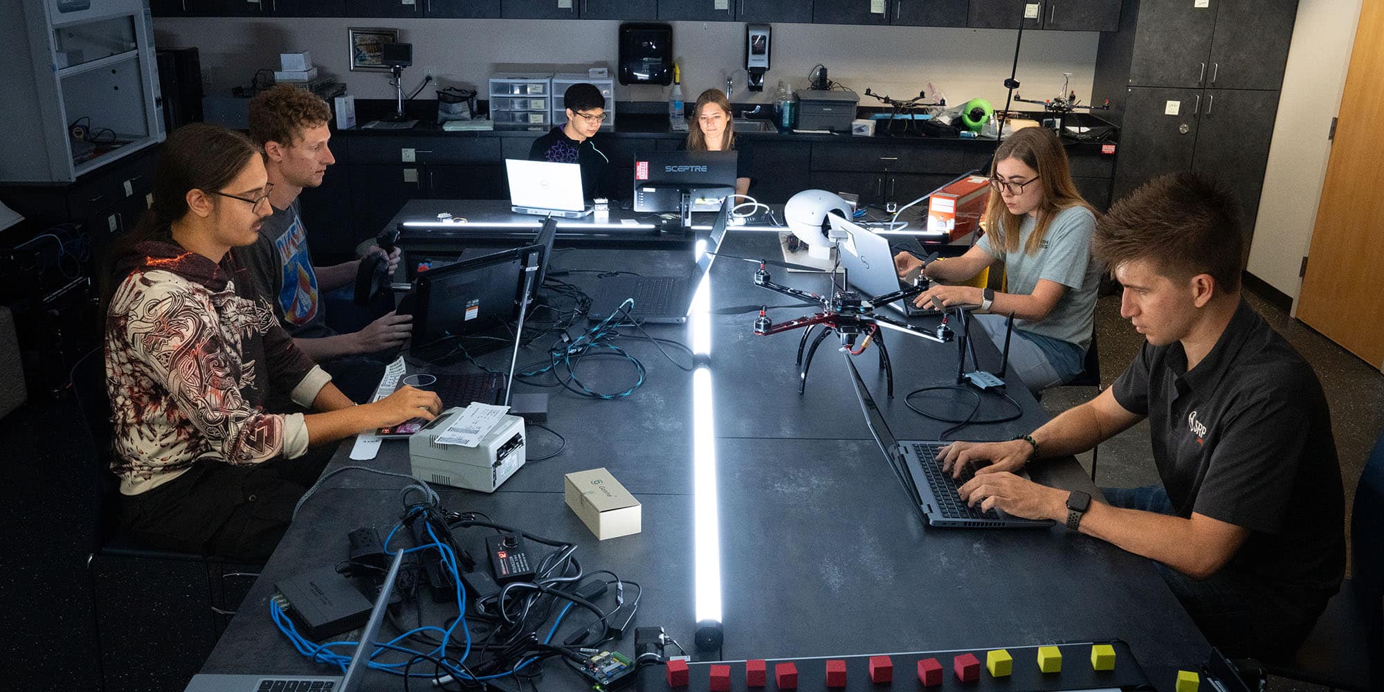 Seven students sit around a large black table, each working on a different laptop. Other equipment, a drone, and several small yellow and red cubes are also on the table.