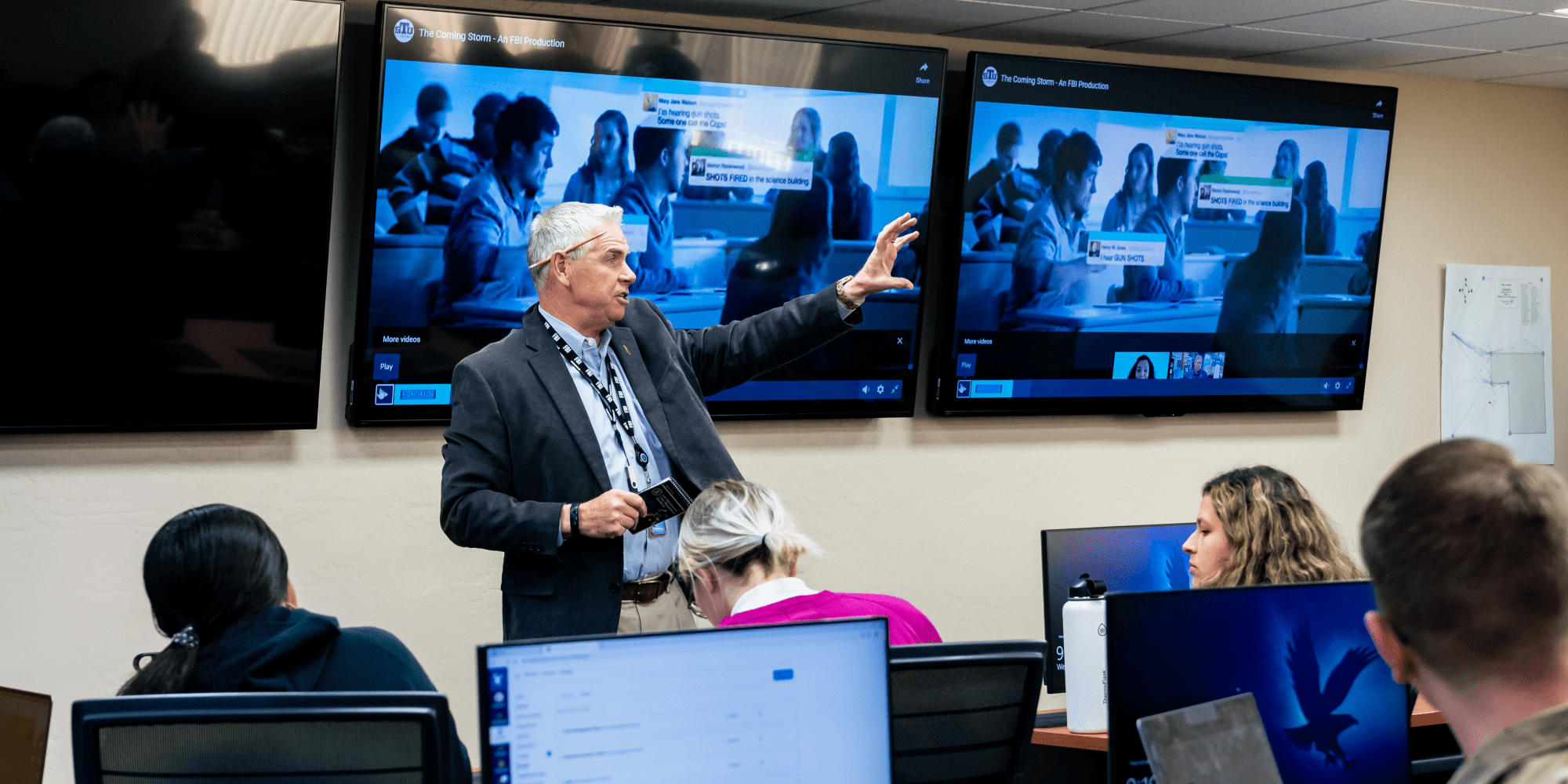 Professor Hooper, a white man with silver hair wearing a suit coat over a button down shirt open at the neck, gestures in front of a classroom. A zoom meeting is displayed on two large monitors behind him.