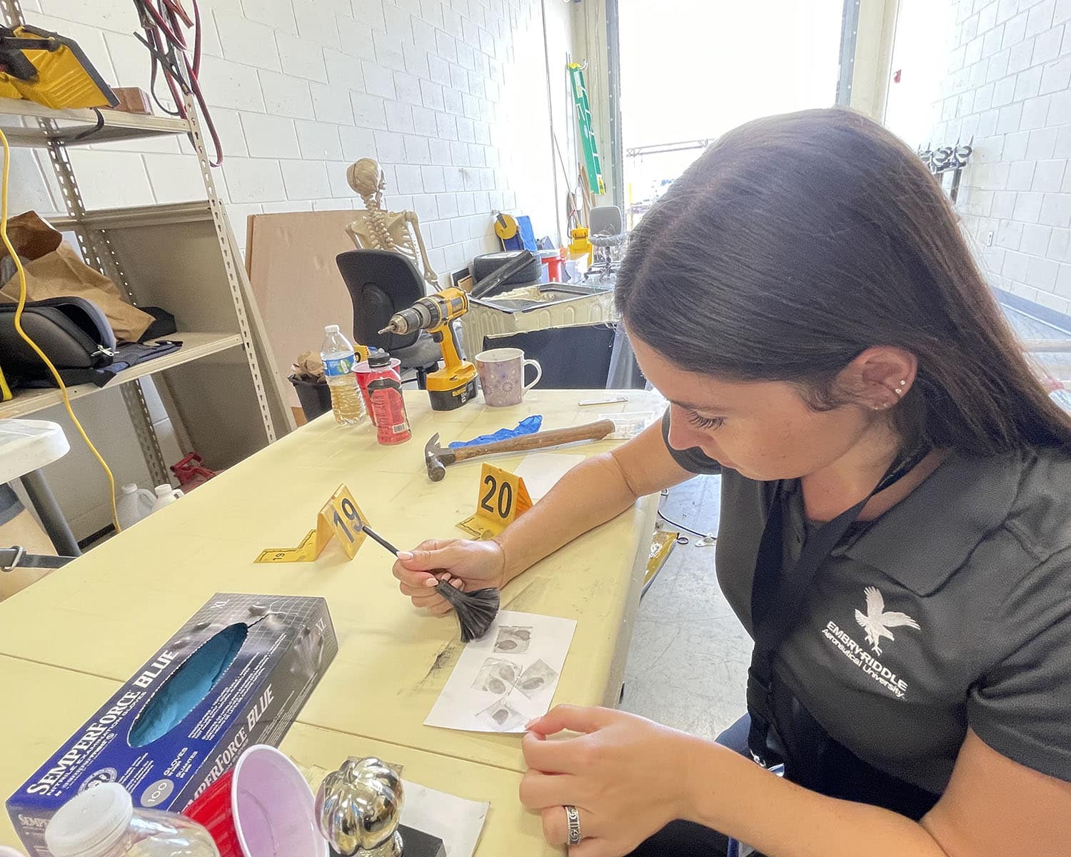 Hannah sits at a table in an evidence room holding a brush over a piece of paper with fingerprints. There's a life-sized plastic skeleton in a chair behind her.