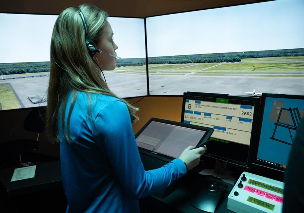 A student wears a headset and monitors several computer screens while watching a simulated airport on three other screens.