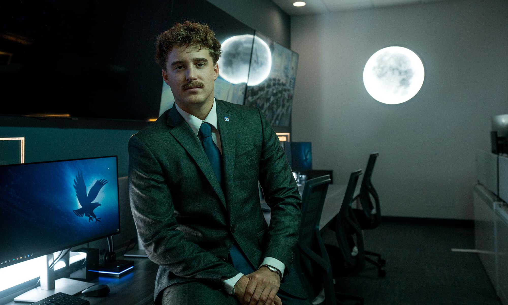 A student with a mustache wearing a suit and tie leans casually against a desk with several computer monitors in a darkened room. A glowing moon shines on the wall behind him.