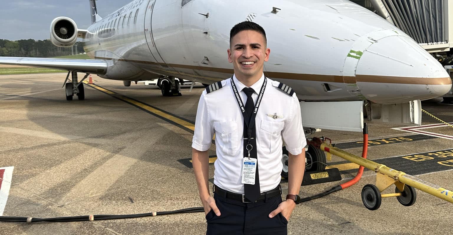 Juan, a man with medium skin tone, wears a pilot uniform and stands in front of a small corporate jet.