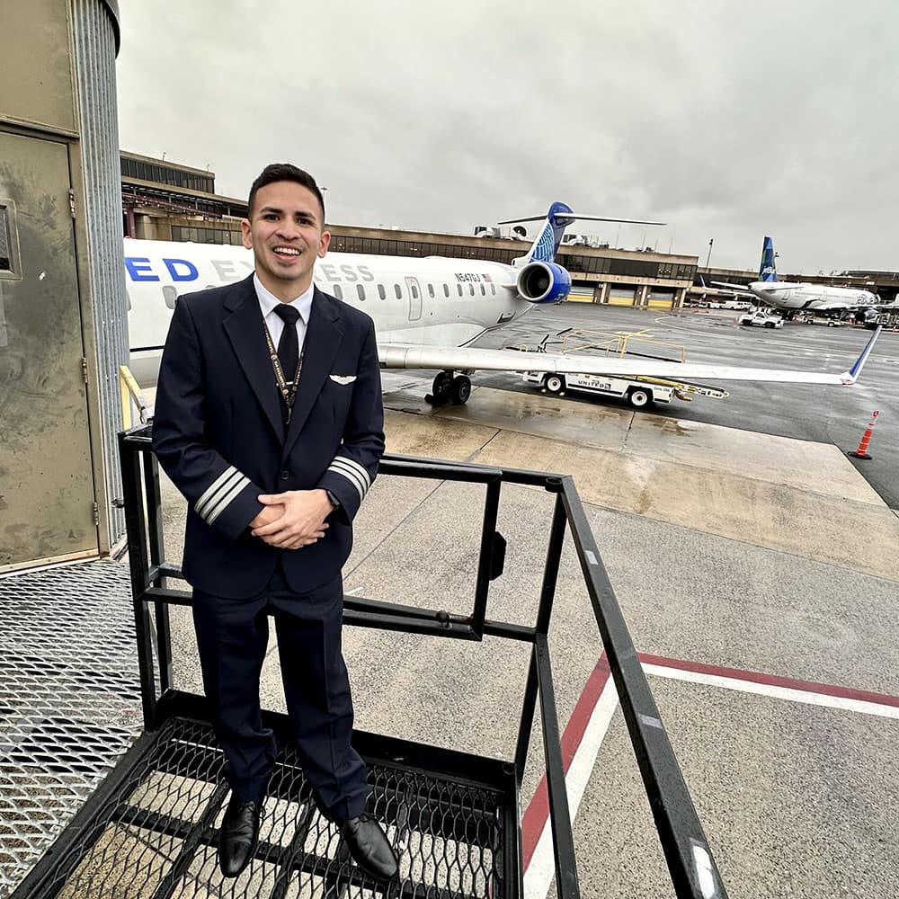 Juan wears a pilot uniform, posing on a staircase with two parked jets behind him.