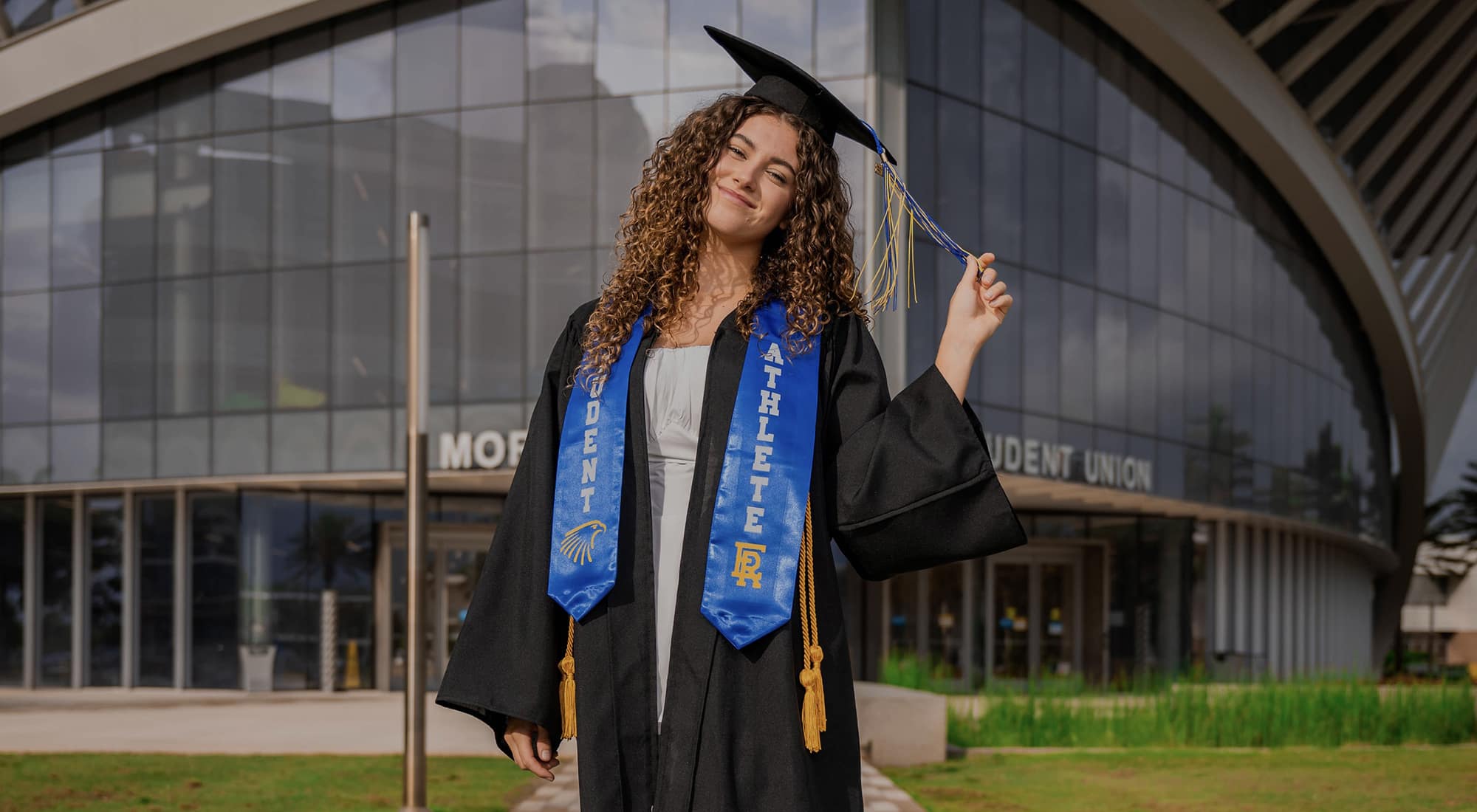 Julia, a white woman with long curly hair, poses with her head tilted, and holding the tassel on her graduation cap with her fingers. She also wears a grad gown, a light blue dress, yellow cords, and a blue sash that reads Student Athlete.