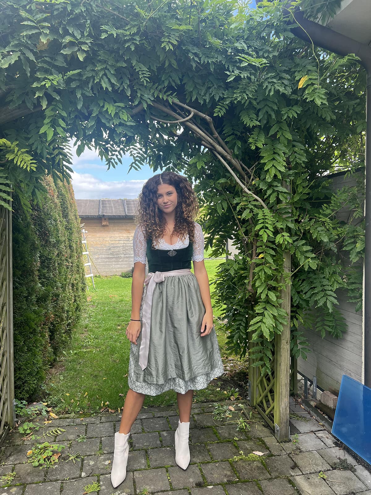 Julia poses in a green dress with a black bodice and white ankle boots, under an archway covered in green vines.