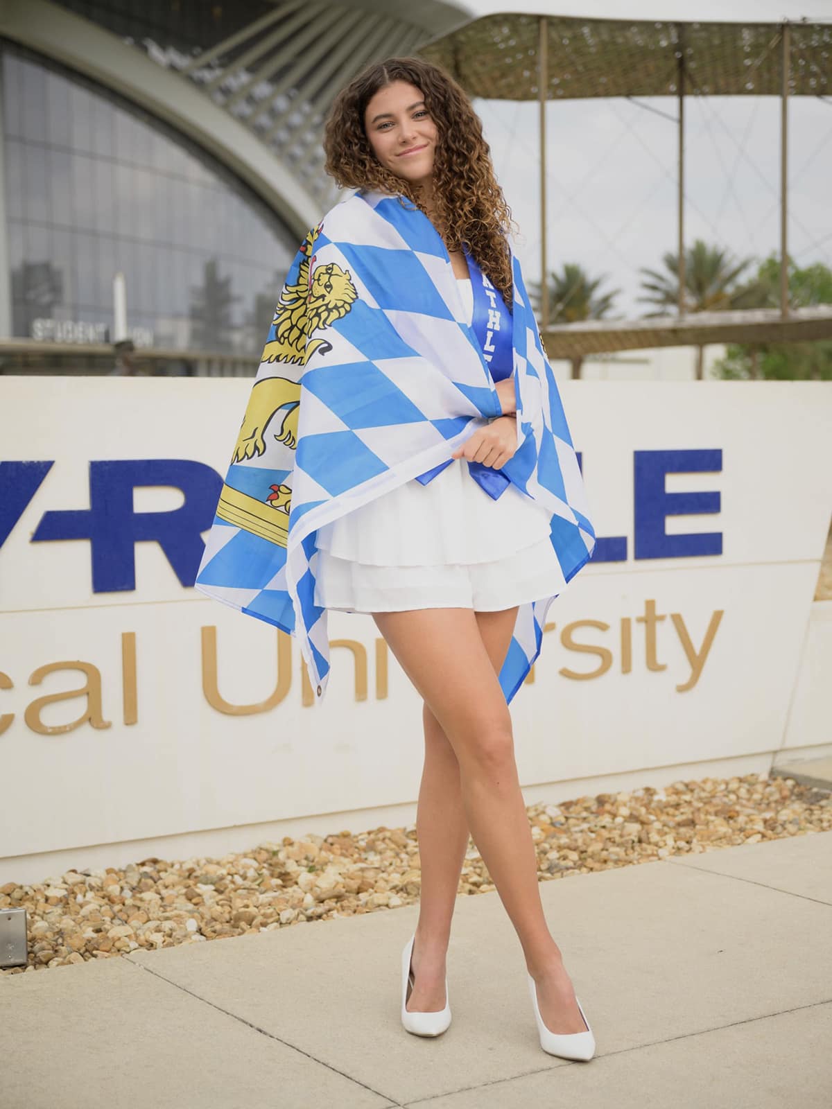 Julia wearing a blue and white flag with a gold lion around her shoulders, a short white skirt, and white pumps.