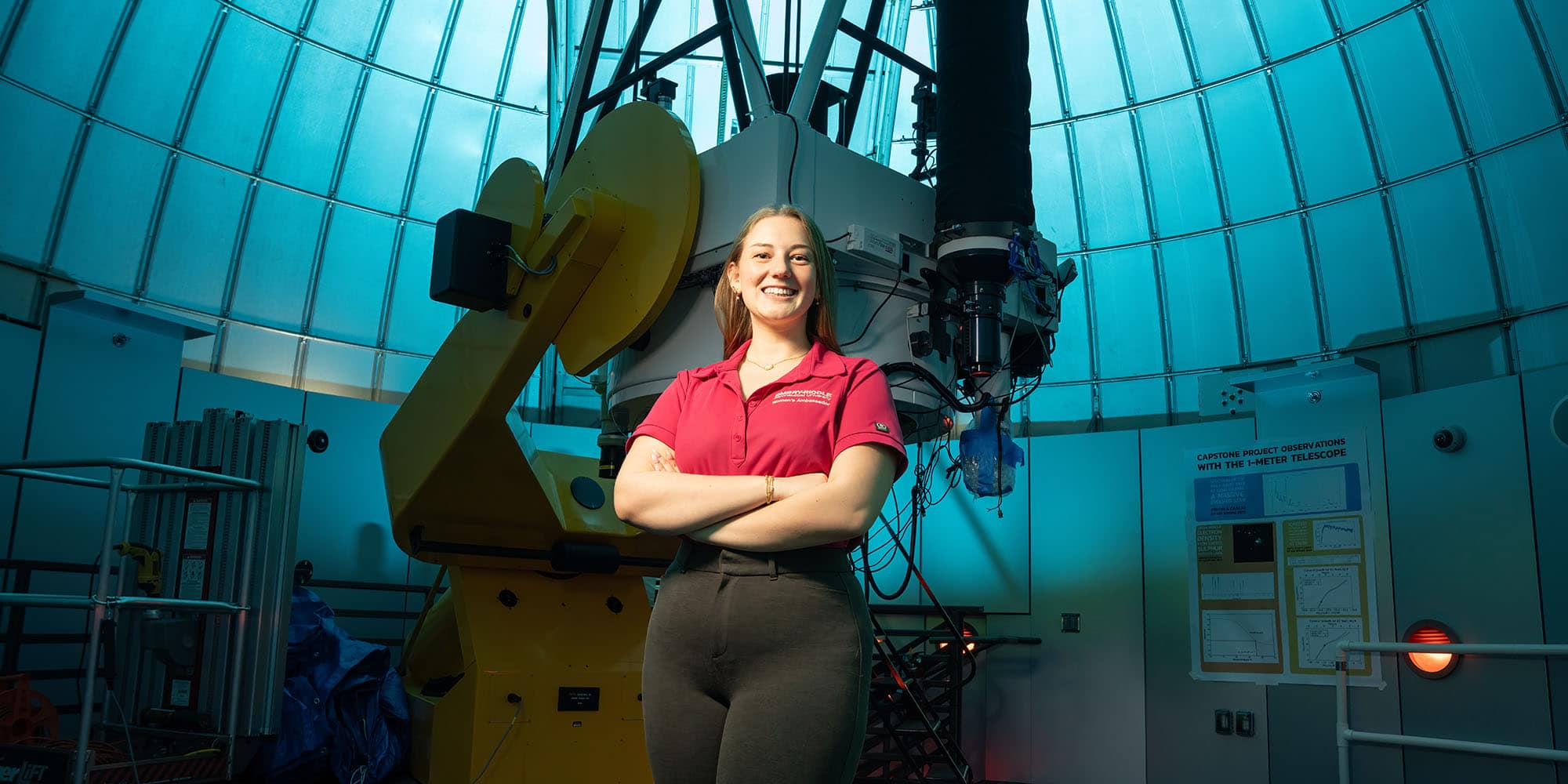 Logan poses with her arms crossed in front of a two-ton telescope; a part of the dome walls visible in the background. Logan is a white woman with dark blond hair pulled back, wearing a red polo shirt with the Embry-Riddle wordmark.