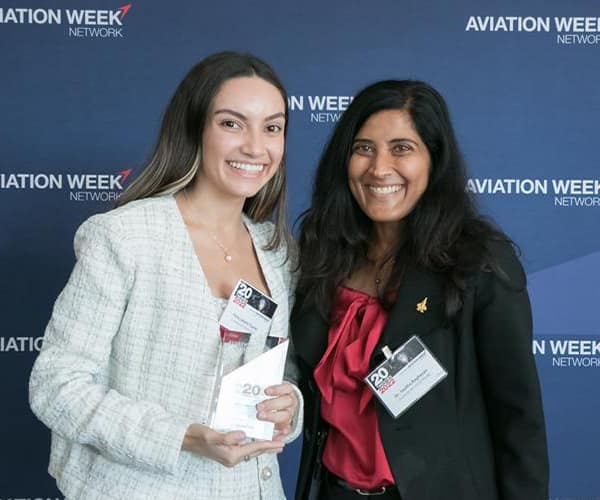 Perla, holding an award, stands with another woman - both are wearing business suits and standing in front of a photo backdrop with the Aviation Week Network logo.