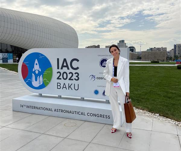 Perla poses in a white pant suit next to a monument sign that reads 74th International Astronautical Congress IAC 2023 Baku.