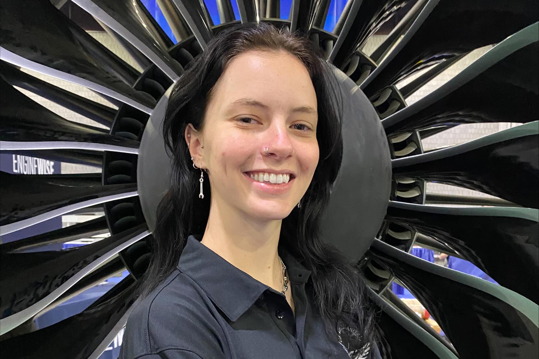 Shelby, a white woman with long dark hair, smiles in front of a large engine fan.
