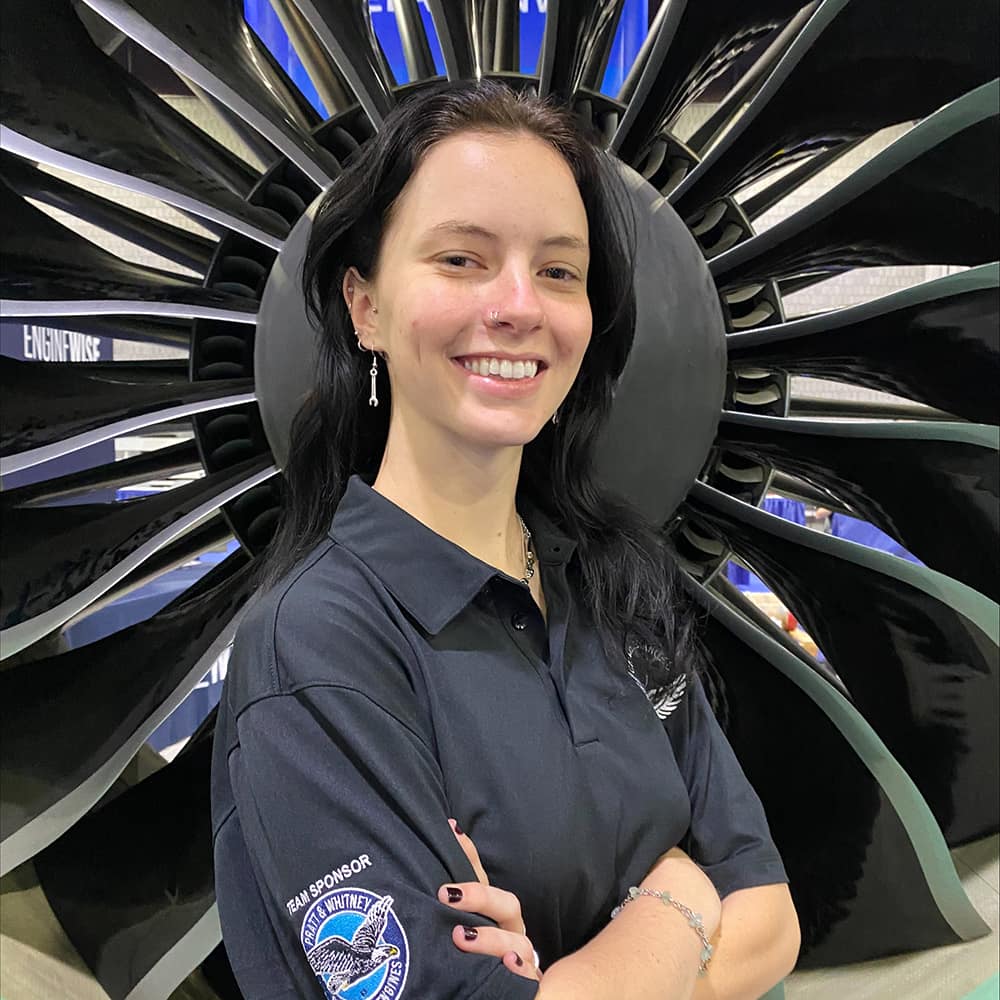 Shelby, a white woman with long dark hair, smiles in front of a large engine fan.