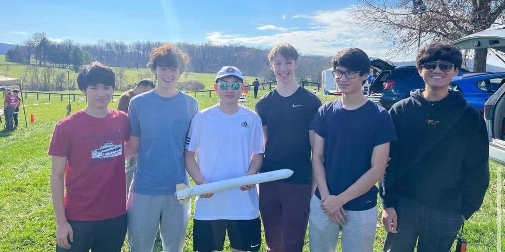 Ryan stands outdoors, holding a small rocket, with five other students, bare autumn trees behind them in the distance