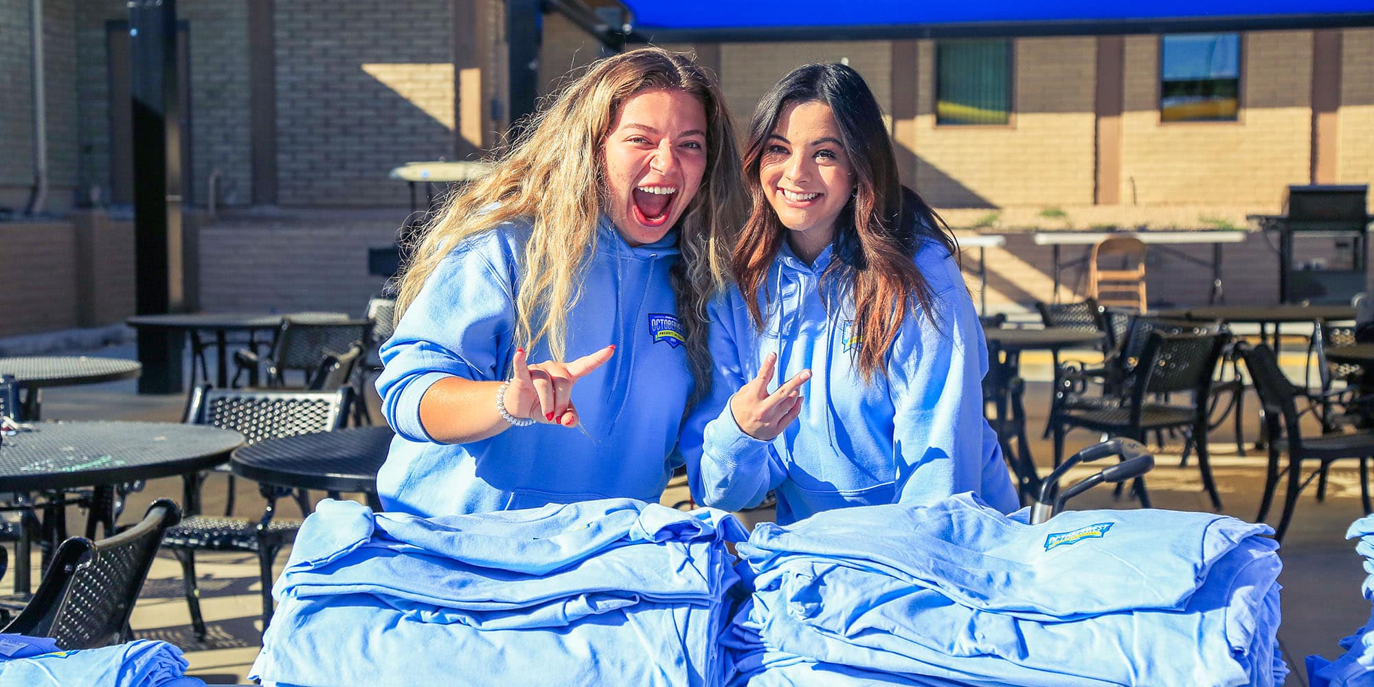 Two students pose with hook 'em and peace hand signals. They're each wearing light blue hoodies with October West logos and stand behind big piles of light blue t-shirts.