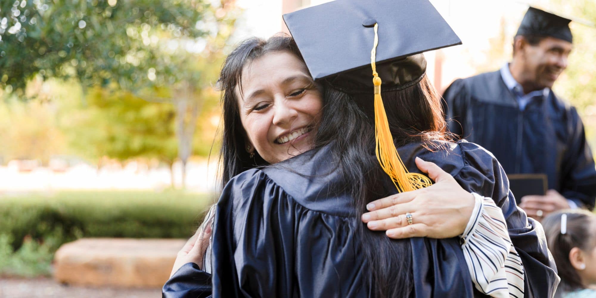 A college student in her graduation gown hugs her smiling mother.