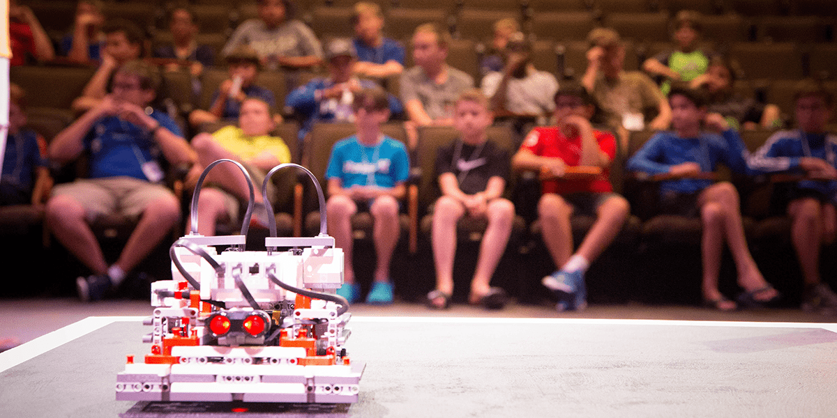 Several middle-school students sit in an auditorium - a small box-shaped robot rests on the stage in the foreground.