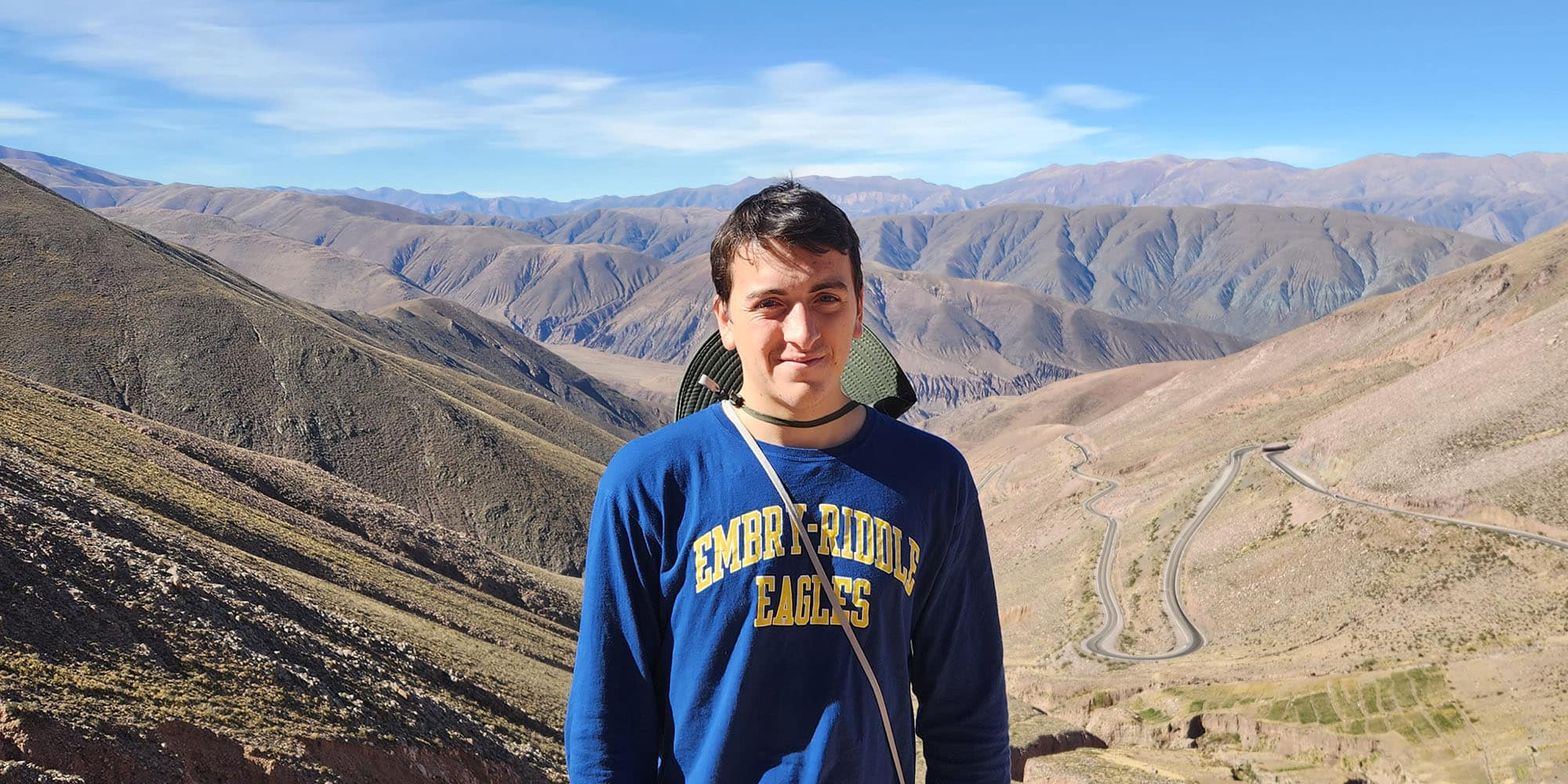 Zackrey poses, squinting into the camera, with a background of brown and gold canyon stretching into the distance. He has light skin tone and is wearing an Embry-Riddle Eagles sweatshirt and a safari hat hanging around his neck.