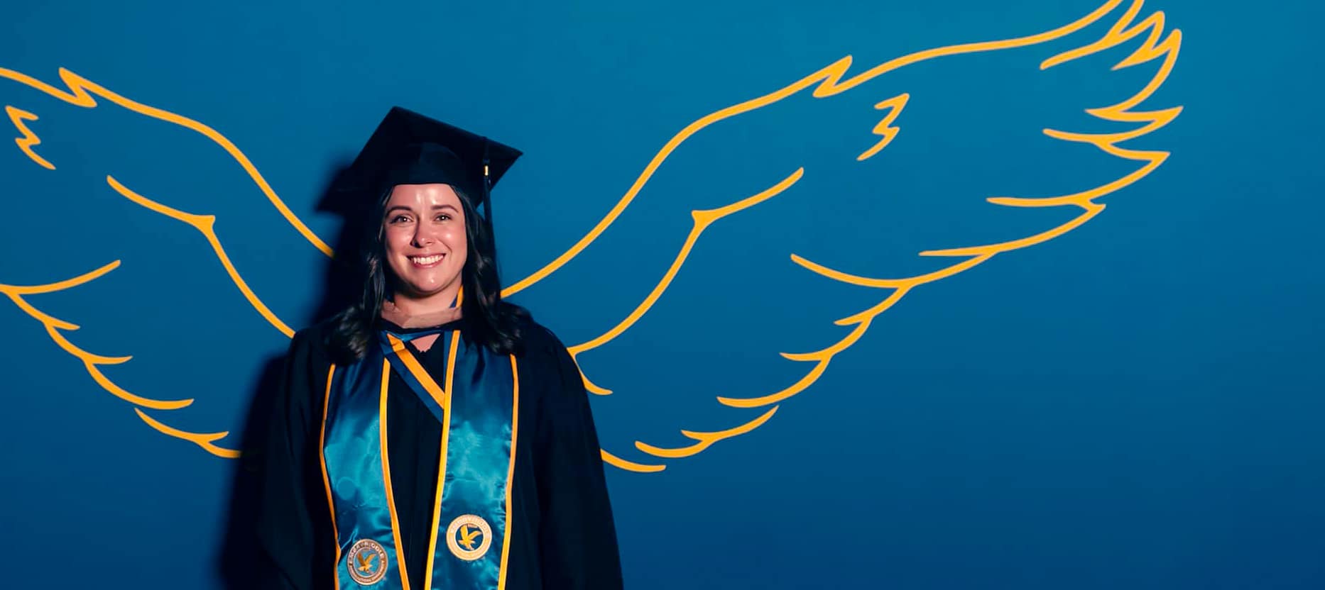 A smiling graduate in cap, gown and sash poses in front of yellow wings printed on a blue wall.