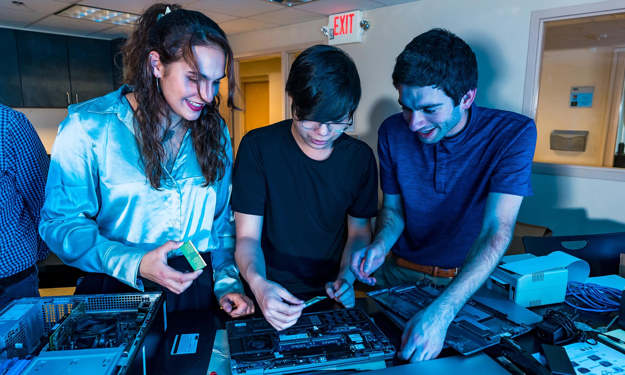 Three students enthusiastically work together on a circuit board in a lab.